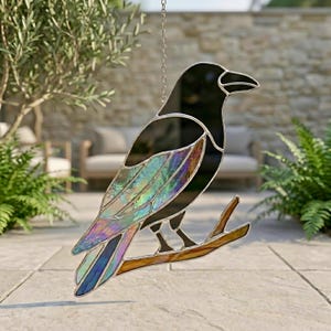 Könnte beinhalten: Ein Glasfenster-Suncatcher mit einem Raben. Der Rabe ist schwarz mit schillernden Flügeln in Blau-, Grün- und Lilatönen. Er sitzt auf einem braunen Ast und hängt an einer silbernen Kette. Der Hintergrund ist eine verschwommene Außenszene.