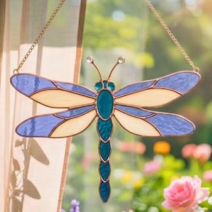 May include: Stained glass dragonfly suncatcher with blue, cream, and teal colors. The dragonfly is suspended by a gold chain. The wings are a mix of blue and cream, and the body is teal. Decorative item for home decor.