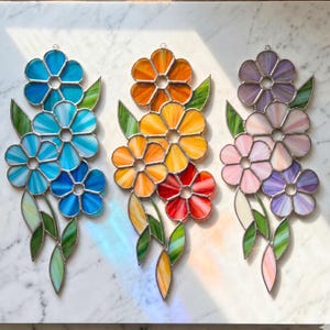 May include: Three vertical stained glass flower arrangements featuring soldered metal frames. The left piece displays blue tones, the centre piece features orange and red hues, and the right piece showcases purple and pink shades with green leaves.