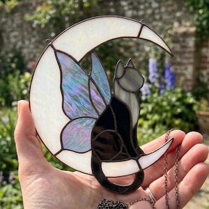 May include: A stained glass suncatcher shaped like a crescent moon with an iridescent wing and a black cat silhouette. The moon is made of opaque white glass, while the cat features dark glass pieces. A metal chain is attached for hanging.