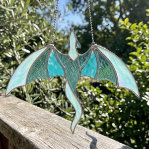 Puede incluir: Atrapasoles de dragón de vidrieras con una cadena plateada para colgar. El dragón presenta una combinación de colores verde azulado y verde con alas iridiscentes. Las alas están muy extendidas y el dragón tiene una cola larga y curvada. El atrapasoles se exhibe al aire libre.