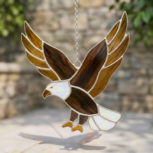 Könnte beinhalten: Glasmalerei-Adler mit ausgebreiteten Flügeln in Braun-, Bernstein- und Weißtönen. Der Adler wird von einer silbernen Kette gehalten, die ein detailliertes Design und Handwerkskunst zeigt. Die Flügel sind weit ausgebreitet und bilden ein dynamisches Dekorationsobjekt.