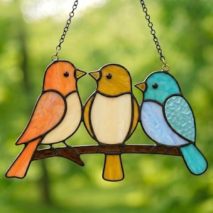 May include: Stained glass art piece featuring three colorful birds perched on a brown branch. The birds are in shades of orange, yellow, and blue, with black outlines and a chain for hanging. A decorative item for home decor.