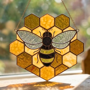 May include: Stained glass bee suncatcher with a honeycomb background. The bee has black and yellow stripes, with translucent wings. The honeycomb is made of yellow and amber glass, and the suncatcher hangs from a gold chain.