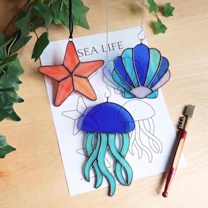 May include: Three stained glass ornaments featuring a five-pointed orange starfish, a blue and teal jellyfish, and a blue and purple seashell. The ornaments are displayed over a paper sheet with the text SEA LIFE and a glass cutting tool.