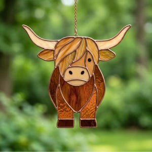 May include: A stained glass Highland cow suncatcher. The cow is made of various shades of brown, tan, and amber glass, with a copper-coloured metal frame. The cow has large horns and a shaggy coat. It is hanging from a gold-coloured chain.