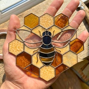 May include: Stained glass bee suncatcher with a honeycomb design. The bee features a black and yellow body and translucent wings. The honeycomb cells are in shades of amber, yellow, and brown. The suncatcher is held in a hand.
