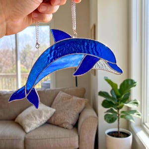 May include: A stained glass dolphin suncatcher featuring deep blue and translucent light blue glass panels held together by silver metal solder lines. The dolphin is suspended by a silver chain and displays a textured, wavy glass pattern.