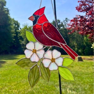 May include: A stained glass suncatcher featuring a bright red cardinal bird perched on a branch with two white flowers and several green leaves. The cardinal has a black face mask and an orange beak. The piece is suspended by a silver metal chain.