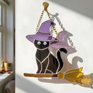 May include: A stained glass suncatcher featuring a black cat wearing a purple witch hat, sitting on a yellow broomstick. The piece is suspended by a gold-toned metal chain and features two white googly eyes attached to the cat's face.