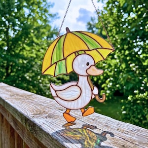 May include: A stained glass suncatcher featuring a white duck with an orange beak and feet, holding a yellow and green umbrella. The piece is constructed with dark metal solder lines and hangs from a thin metal chain, displaying translucent glass.
