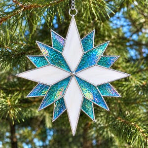 May include: A multi-pointed star ornament constructed from stained glass. The design features alternating opaque white diamond-shaped panels and iridescent, textured teal and blue glass segments, all held together by thin silver metal solder lines.