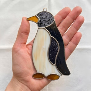 May include: A stained glass penguin ornament featuring a textured black head and back, a smooth cream-coloured belly, and a bright orange beak and feet. The piece is constructed with dark metal solder lines and includes a small loop at the top.