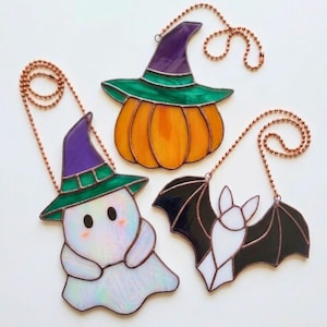 May include: Three stained glass suncatchers featuring a ghost wearing a purple and green witch hat, an orange pumpkin with a purple and green witch hat, and a white bat with black wings, each suspended by a copper-coloured metal ball chain.