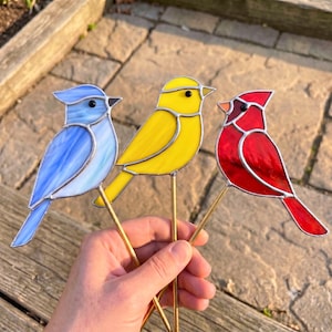 Peut inclure: Trois ornements d'oiseaux en vitrail sur des tiges métalliques. Un oiseau est bleu clair avec des stries blanches, un est jaune vif et un est rouge éclatant. Chaque oiseau possède une petite perle noire pour l'œil et des lignes de soudure argentées.