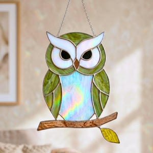 May include: A stained glass owl suncatcher featuring iridescent white eyes, a light blue iridescent chest, and olive green wings. The owl is perched on a textured brown glass branch with a single yellow leaf, suspended by a silver metal chain.