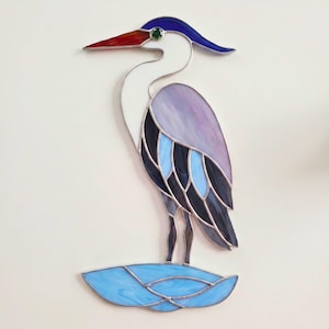 May include: A stained glass suncatcher shaped like a heron standing on a blue water base. The bird features a white neck, a dark blue crest, a red beak with a green eye accent, and a body composed of light purple, light blue, and dark blue glass.
