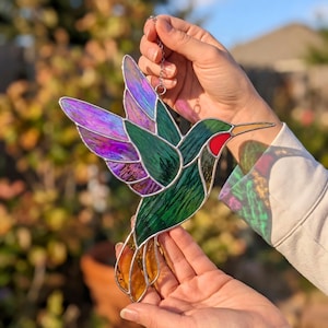 Op de afbeelding: Gebrandschilderd glazen kolibrie zonnevanger met iriserende paarse en groene vleugels, een rode keel en een oranje snavel. De decoratieve vogel wordt aan een zilveren ketting gehouden, een uniek kunstwerk.