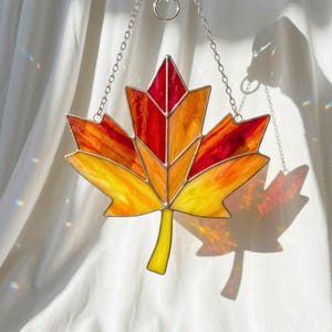 May include: Stained glass maple leaf in shades of red, orange, and yellow, with a silver chain for hanging. The leaf design is reminiscent of autumn foliage. A decorative piece for home decor.