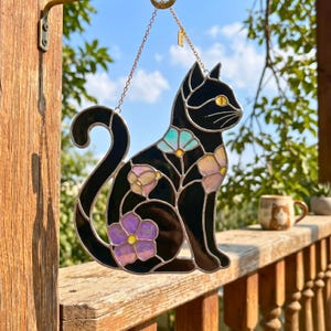 Pode incluir: Um apanhador de sol de vitral em forma de gato sentado, com painéis de vidro preto e detalhes florais iridescentes em tons de roxo, rosa e azul. O gato tem um olho de vidro amarelo e está suspenso por uma fina corrente de metal.