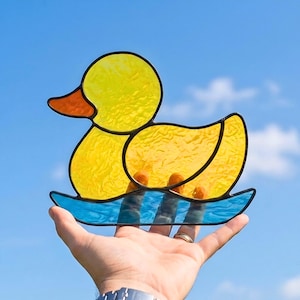 May include: A stained glass duck with yellow body and head, orange beak, and blue base. The duck is held up against a blue sky. The glass has a textured appearance, and the duck is outlined in black.