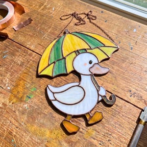 Op de afbeelding: Een glas-in-lood suncatcher met een witte eend met oranje voeten en een lichtoranje snavel, die een gele en groene paraplu vasthoudt. Het werk is gemaakt met donkere metalen soldeerlijnen en bevat een koperkleurige metalen ophangketting.