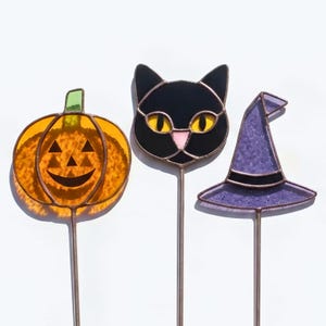 Puede incluir: Tres estacas de jardín de vidrio tintado que incluyen una calabaza naranja con tallo verde, una cabeza de gato negro con ojos amarillos y nariz rosa, y un sombrero de bruja morado, unidos a varillas de metal delgadas para su exhibición.