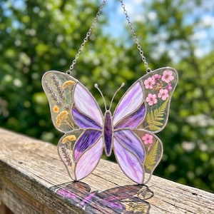 Può includere: Un acchiappasole a forma di farfalla con pannelli di vetro dai motivi viola e bianchi incorniciati in metallo. Le ali contengono fiori secchi pressati, tra cui lavanda, rametti gialli, fiori rosa e foglie di felce verdi, appeso a una catena.