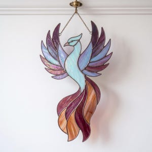 Può includere: Un acchiappasole in vetro colorato a forma di fenice con le ali spiegate. L'uccello presenta un corpo azzurro, piume delle ali viola e iridescenti e una coda composta da segmenti di vetro arancione, ambra e viola scuro con saldature.