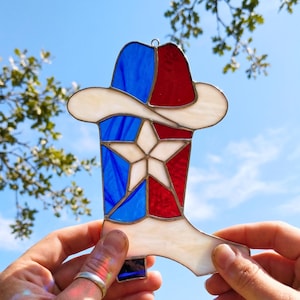 Puede incluir: Un atrapasoles de vidrieras con forma de bota de vaquero con un sombrero encima. El diseño presenta una estrella blanca central, con secciones de vidrio translúcido azul, rojo y crema unidas por líneas de soldadura de metal oscuro.