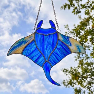 May include: A stained glass suncatcher shaped like a manta ray, featuring vibrant blue marbled glass in the center and wings, with cream and light blue swirled glass accents on the wing tips. It hangs from a silver-toned metal chain.