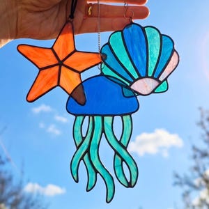 Puede incluir: Tres atrapasoles de vidrio tintado que presentan una estrella de mar naranja brillante, una medusa azul y turquesa con largos tentáculos fluidos y una concha festoneada en tonos azules y blancos, unidos por finas cadenas de metal.