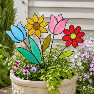 May include: A stained glass garden stake featuring four distinct flowers: a blue tulip, a yellow sunflower with a textured center, a pink tulip, and a red daisy with a yellow center, all connected by thin black metal stems and green leaves.