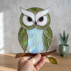 Puede incluir: Un atrapasoles de vidriera con forma de búho, compuesto por paneles de vidrio blanco y verde iridiscente unidos por líneas de soldadura de metal plateado. El búho está posado sobre una rama de vidrio marrón con una sola hoja verde.