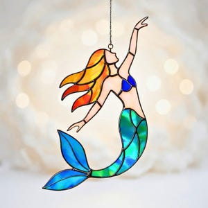 Può includere: Suncatcher a forma di sirena in vetro colorato. La sirena ha capelli arancioni e gialli, un top blu e una coda verde e blu. Il suncatcher è sospeso da una catena.