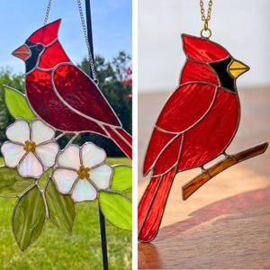 May include: Stained glass cardinal bird suncatcher. The red bird has a black mask and yellow beak. The bird is perched on a branch or next to white flowers with green leaves. The suncatcher hangs from a chain.