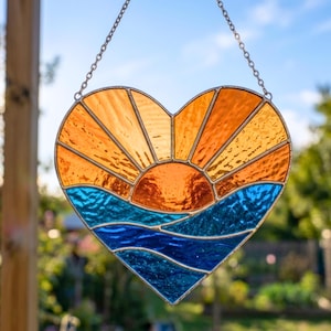 May include: Heart-shaped stained glass suncatcher with a sunset design. The sun is orange, with orange and yellow rays. The ocean is blue and the sky is a light blue. The suncatcher hangs from a silver chain.