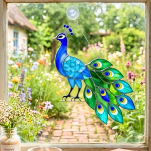 Puede incluir: Un adorno de ventana de pavo real de vidrieras con un cuerpo azul vibrante y un abanico de plumas de cola verdes, azules y amarillas. El pavo real está suspendido frente a una ventana con vistas al jardín.