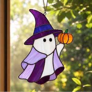 Op de afbeelding: Een glas-in-lood suncatcher in de vorm van een geest met een paarse heksenhoed en een lichtpaarse cape. De geest heeft twee zwarte ronde ogen en houdt een kleine oranje pompoen met een groene steel vast. Het hangt aan een dunne metalen ketting.