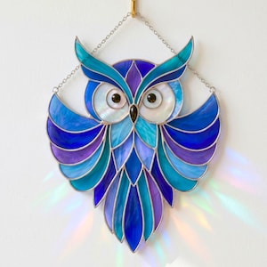 May include: A stained glass owl suncatcher featuring a vibrant palette of deep blue, turquoise, and purple glass segments. The owl has two large, circular white iridescent eyes with black centers, all held together by silver metal solder lines.