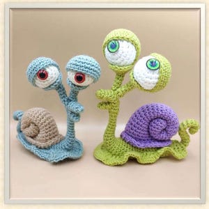 May include: Two whimsical, handmade crochet snails. One has a blue body, brown shell, and two eyes with red irises. The other has a green body, purple shell, and three eyes with green irises. The snails are on a neutral background.