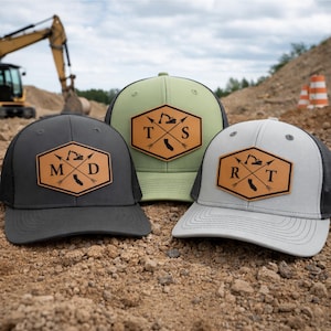 May include: Three trucker hats with a leather patch featuring a construction vehicle, arrows, and the California outline. The hats are in gray, olive green, and light gray with black mesh backs. The patches have the letters MD, TS, and RT.