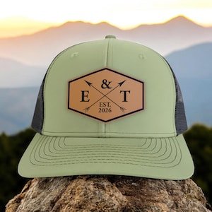 May include: A sage green trucker hat with a black mesh back. The hat features a brown leather patch with the letters "E & T" and the year "2026" in a hexagon shape. The hat is sitting on a rock.