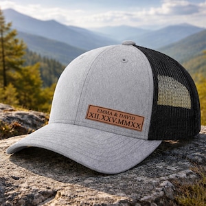 May include: A gray baseball cap with a black mesh back. The cap features a leather patch with the names "EMMA & DAVID" and the date "XII.XXV.MMXX" in Roman numerals. The cap is sitting on a rock.