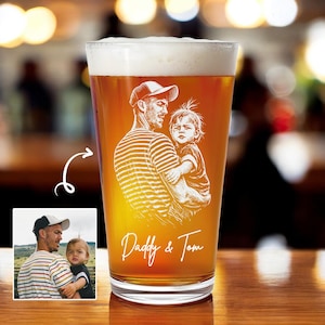 May include: A pint glass filled with beer, featuring a custom etched design of a father holding a child. The design includes the text "Daddy & Tom" in a script font. The glass has a foamy head and is set against a blurred background.