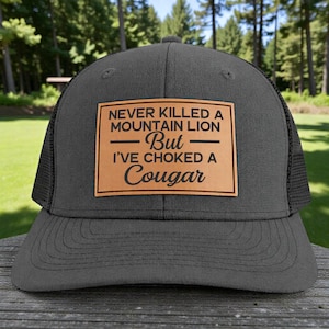 May include: A charcoal gray trucker hat with a black mesh back and a curved brim. The hat features a rectangular, tan leather patch with the text "NEVER KILLED A MOUNTAIN LION - But - I'VE CHOKED A Cougar" in black.
