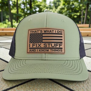 May include: A green and black trucker hat with a brown leather patch. The patch features the American flag and the text "THAT'S WHAT I DO, I FIX STUFF, AND I KNOW THINGS."