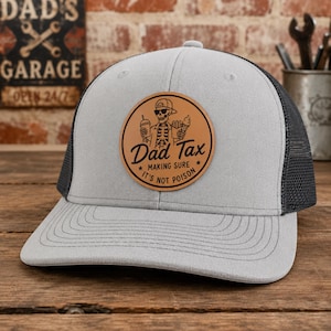 May include: A gray trucker hat with a black mesh back and a curved brim. The hat features a circular brown leather patch with the words "Dad Tax" and a skeleton graphic holding food and a drink. The text "Making sure it's not poison" is also on the patch.