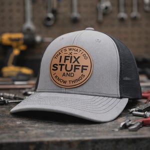 May include: A gray and black trucker hat with a circular leather patch. The patch features the text "THAT'S WHAT I DO, I FIX STUFF AND I KNOW THINGS" in a circular design. The hat has a curved brim and a mesh back.