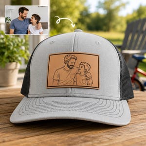 May include: Gray trucker hat with a black mesh back and a light brown leather patch. The patch features a black line drawing of a man and a boy embracing. The hat is sitting on a wooden surface.